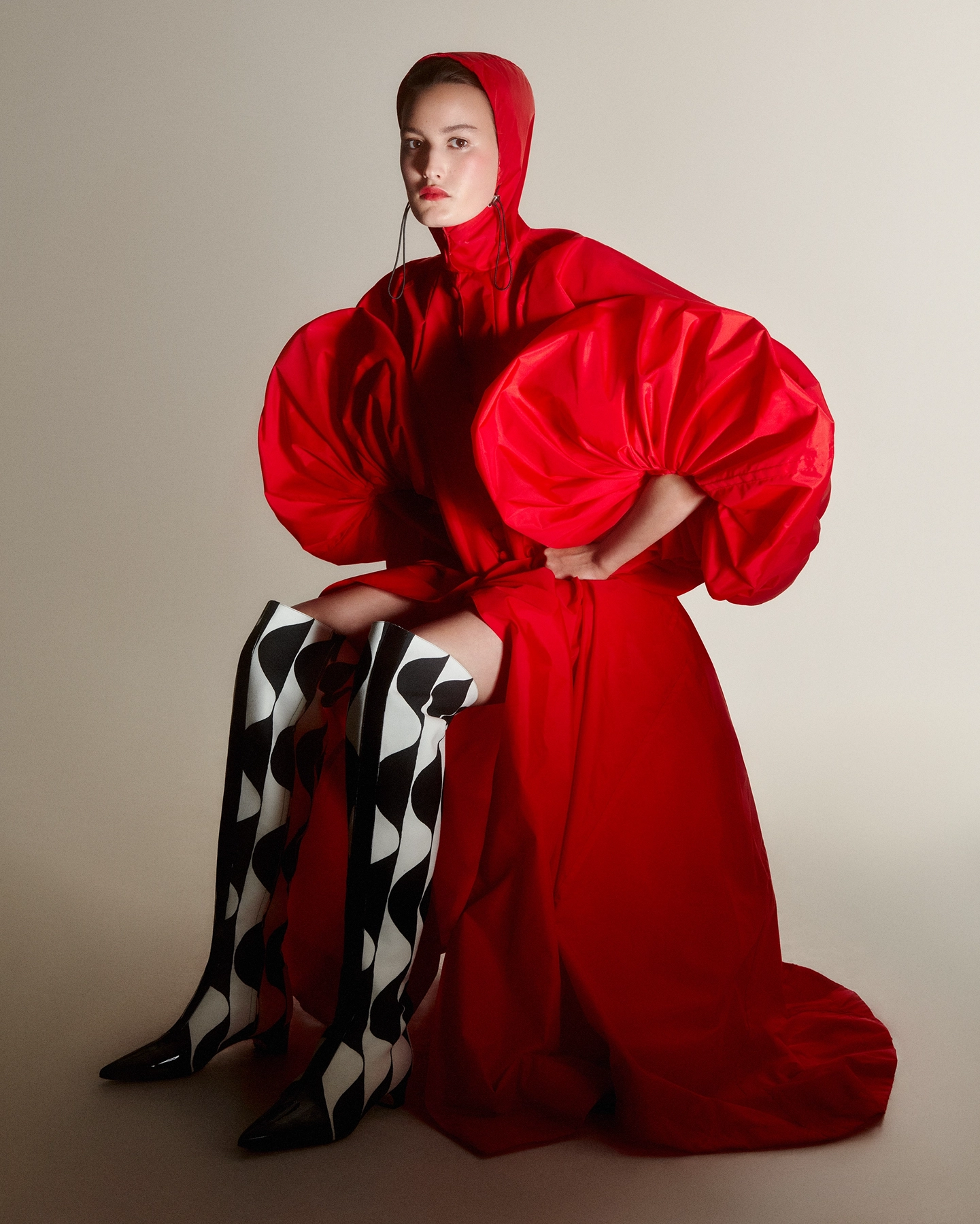 Seated model in voluminous red hooded gown with harlequin-print knee-high boots for Harpers Bazaar Arabia