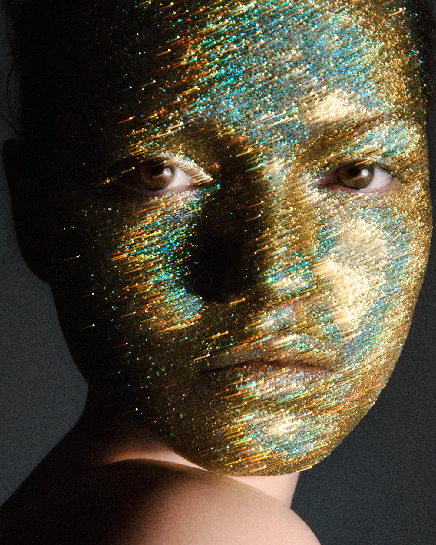 Close-up with iridescent gold and teal glitter covering entire face, direct gaze through textured mask
