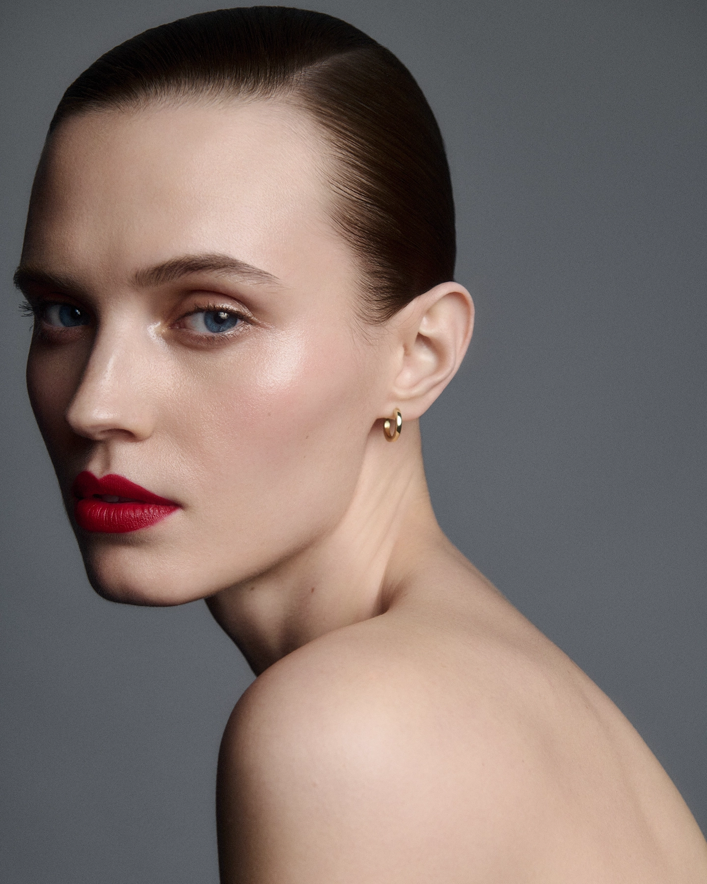 Agnes three-quarter beauty portrait with slicked hair, red lip and gold hoop earring on grey backdrop