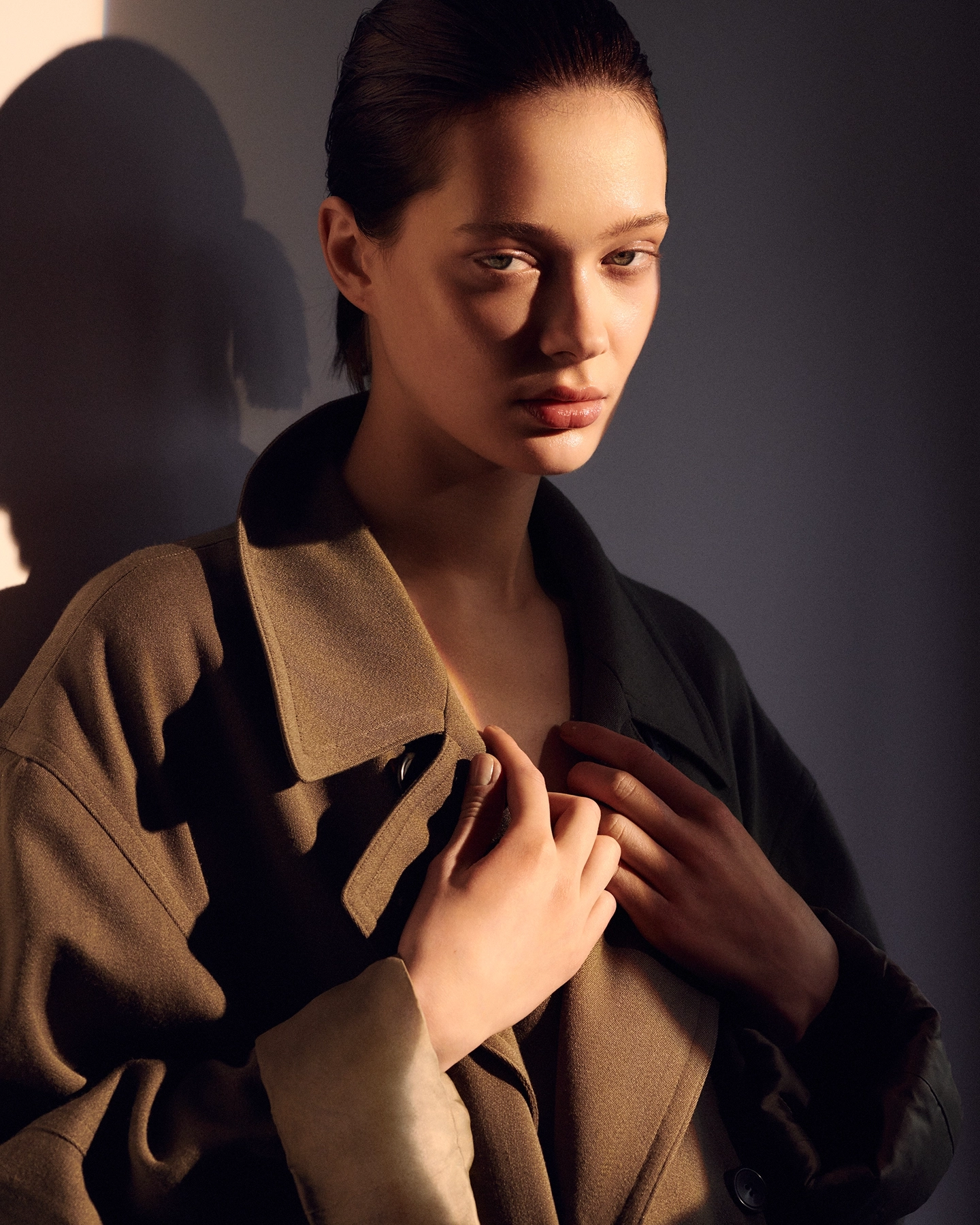 Tanya in olive two-tone coat clutched at the collar with warm directional light casting shadow on studio wall