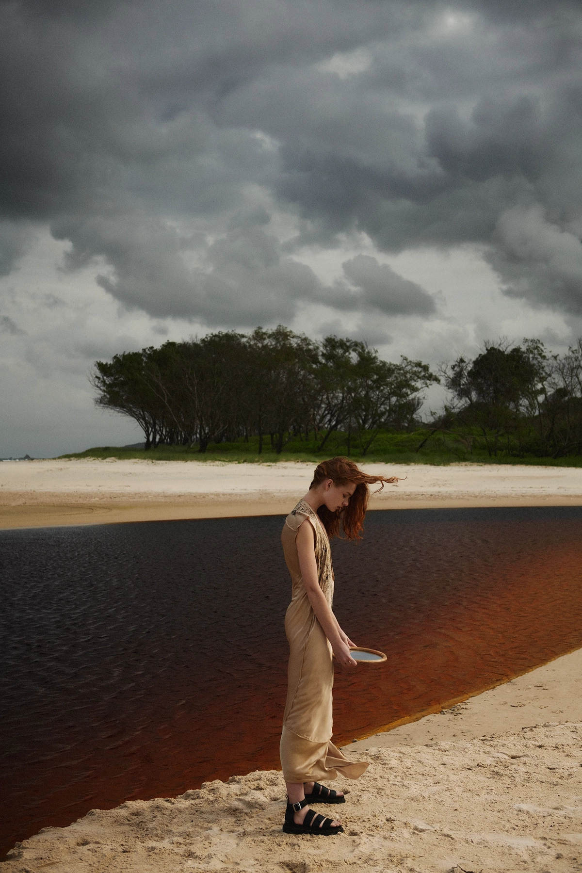 Model standing at waters edge in linen dress under dramatic storm clouds with windswept hair for The Paradiso Magazine