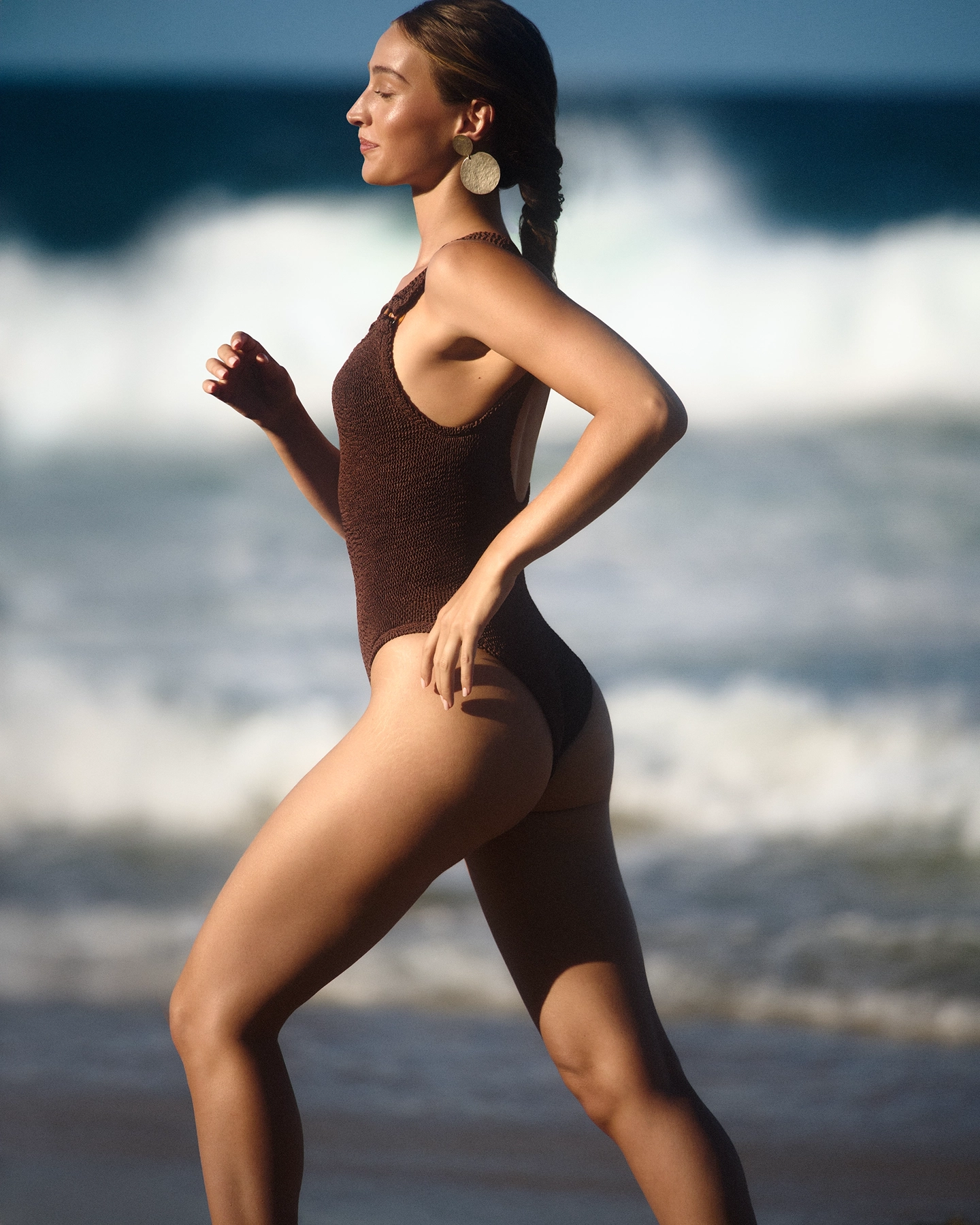 Gabrielle in brown textured swimsuit walking on beach in profile against breaking waves for 5 Eleven Magazine