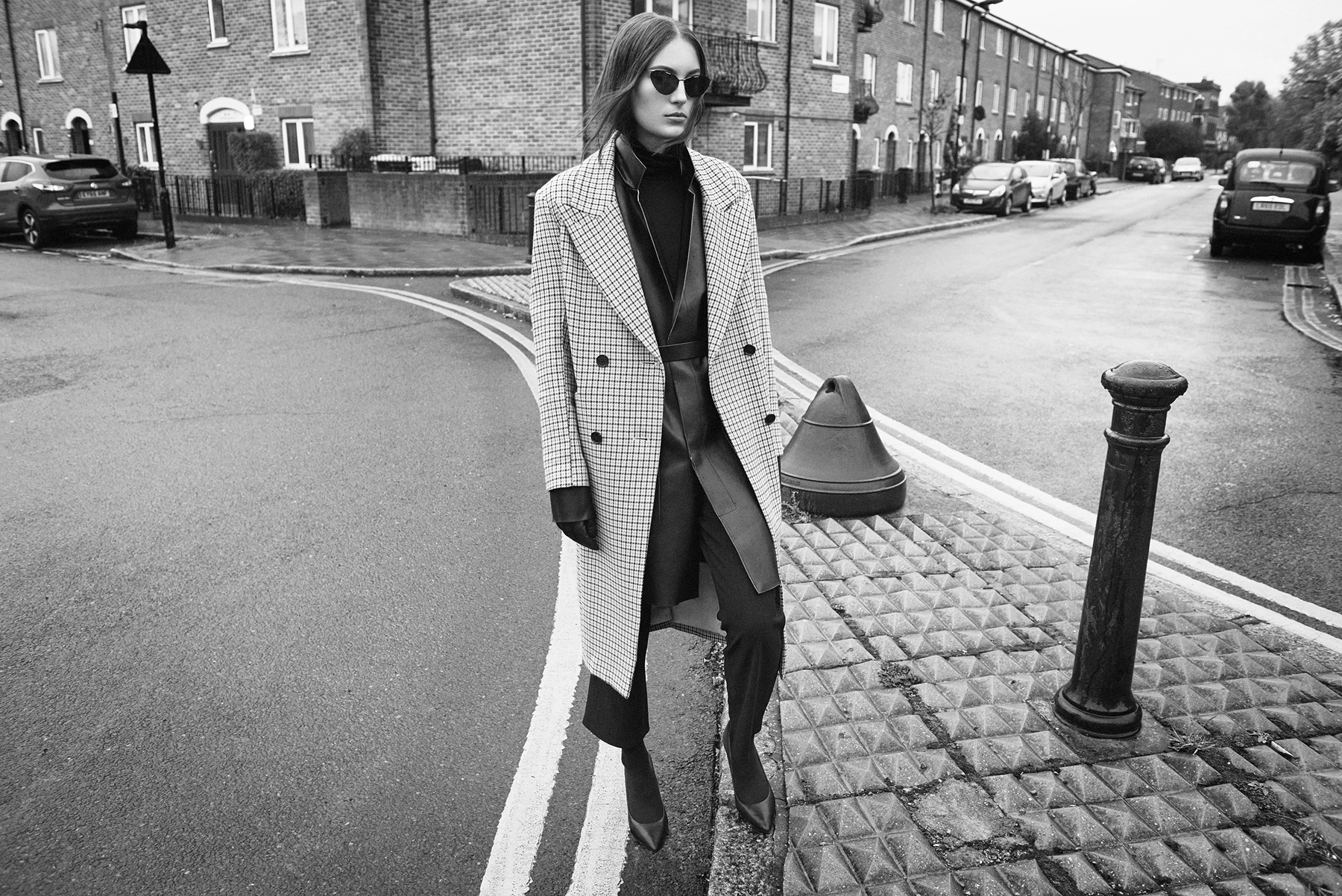 Black and white street fashion shot of model in houndstooth overcoat crossing a rainy London street