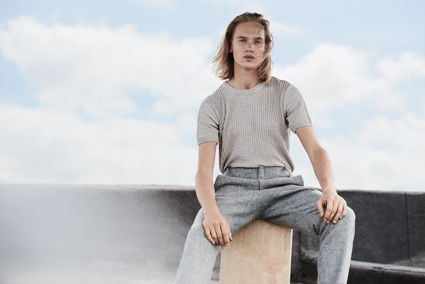 Marcel in grey ribbed knit and tailored trousers seated on rooftop ledge against cloudy sky for Male Model Scene, styled by Carlos Mangubat
